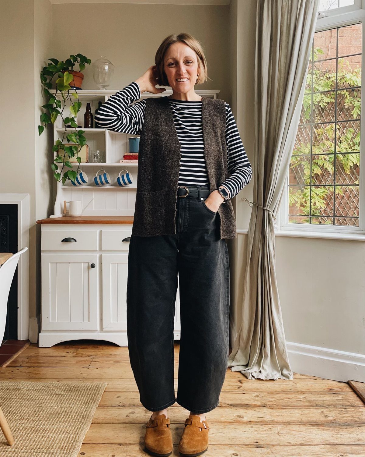 Woman wearing a dark vest, striped shirt, wide-leg black trousers, and brown Birkenstock clogs.