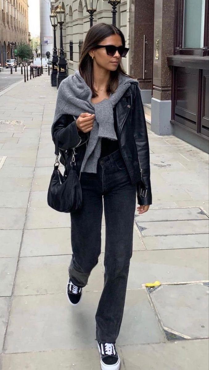A woman walking down a city street wearing black leather jacket, black jeans, and black sneakers.