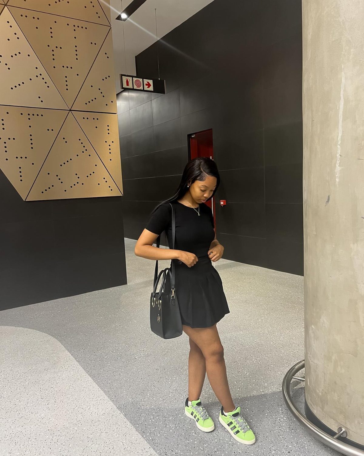A woman in a black mini dress and lime green sneakers posing in a hallway.