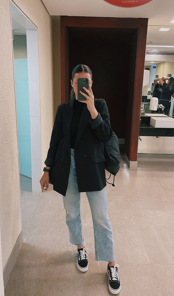 Woman in a black blazer, light blue jeans, and black sneakers posing for a mirror selfie.