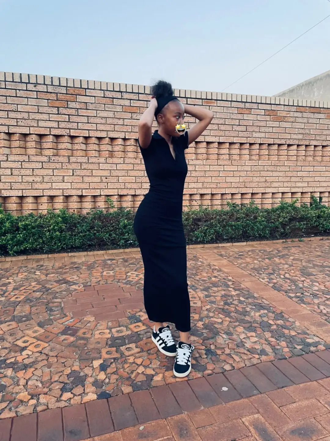 A woman wearing a long black dress and black sneakers standing on a brick patio.