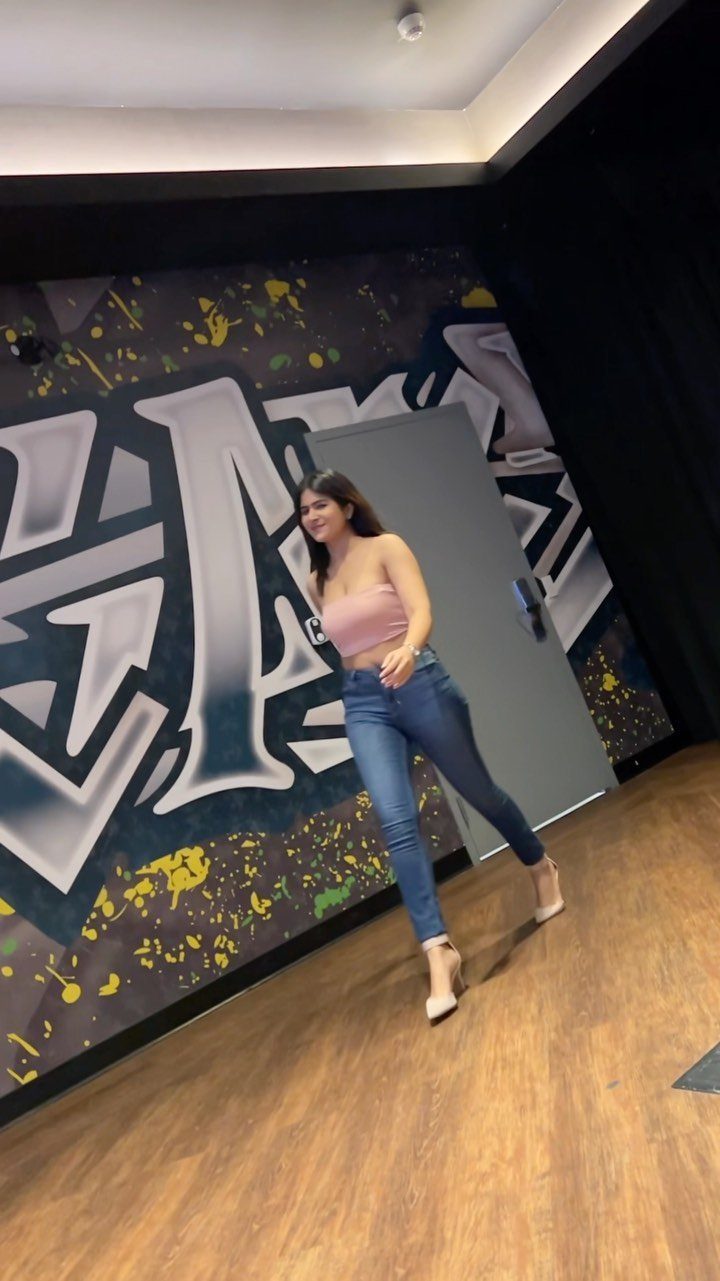 A woman wearing a pink top, blue jeans, and beige high heels posing against graffiti.