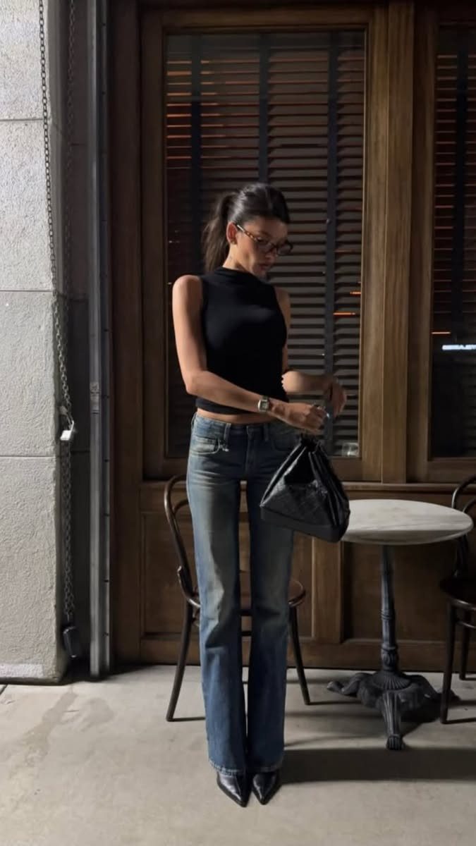 A woman wearing a black sleeveless turtleneck, blue jeans, and black high heels standing outdoors.