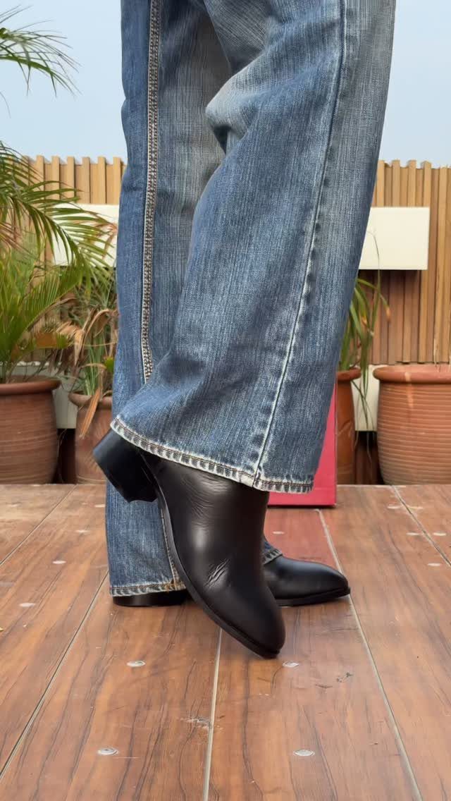Person wearing blue denim jeans and black leather Chelsea boots standing on a wooden deck.