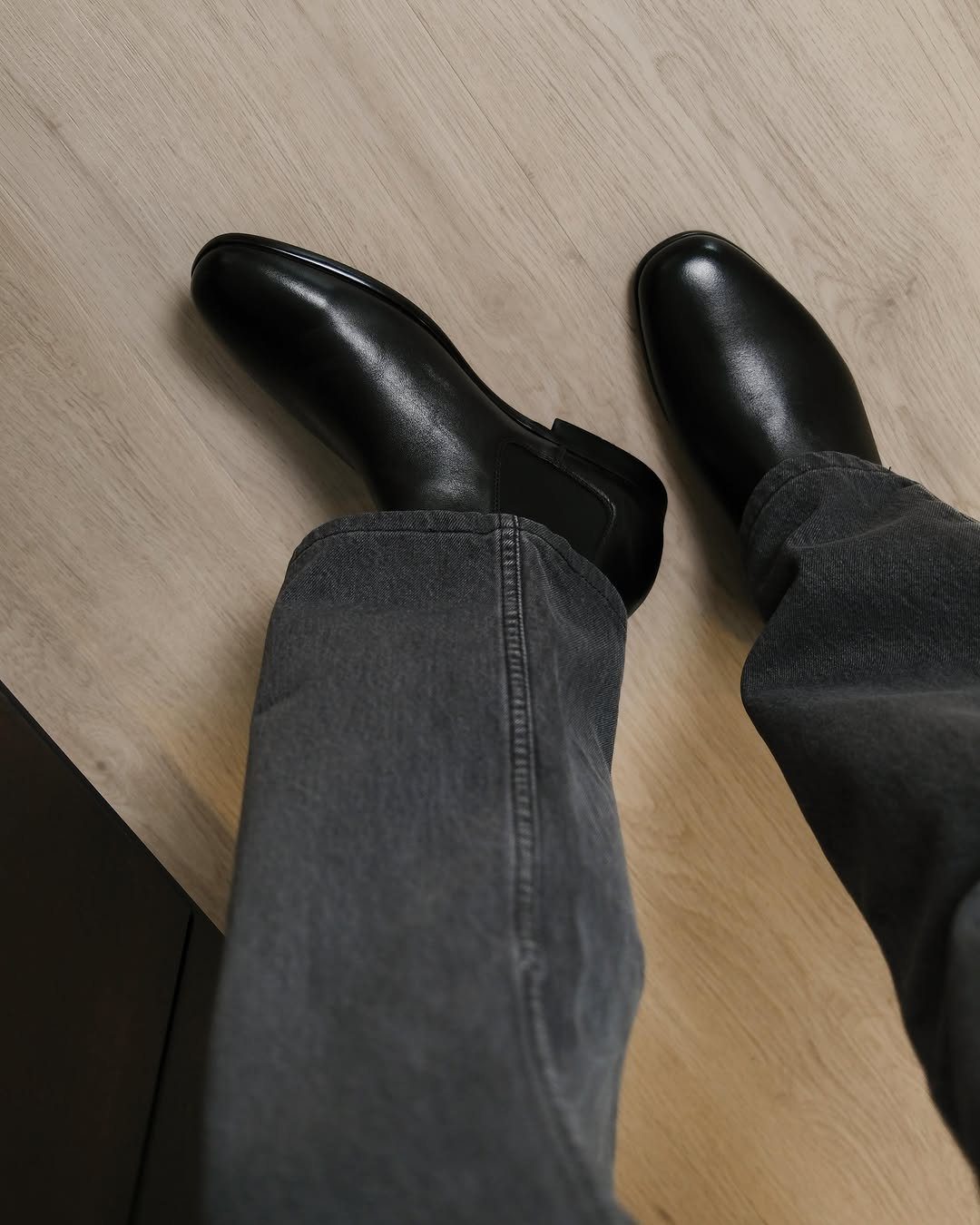 Black leather Chelsea boots paired with dark grey denim jeans on a light wood floor.