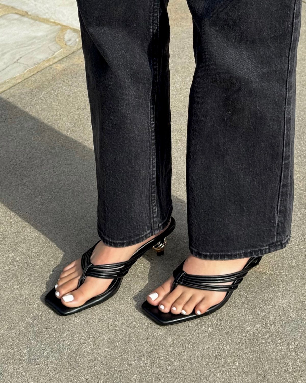 Person wearing black denim jeans and black thong kitten heel sandals on pavement.