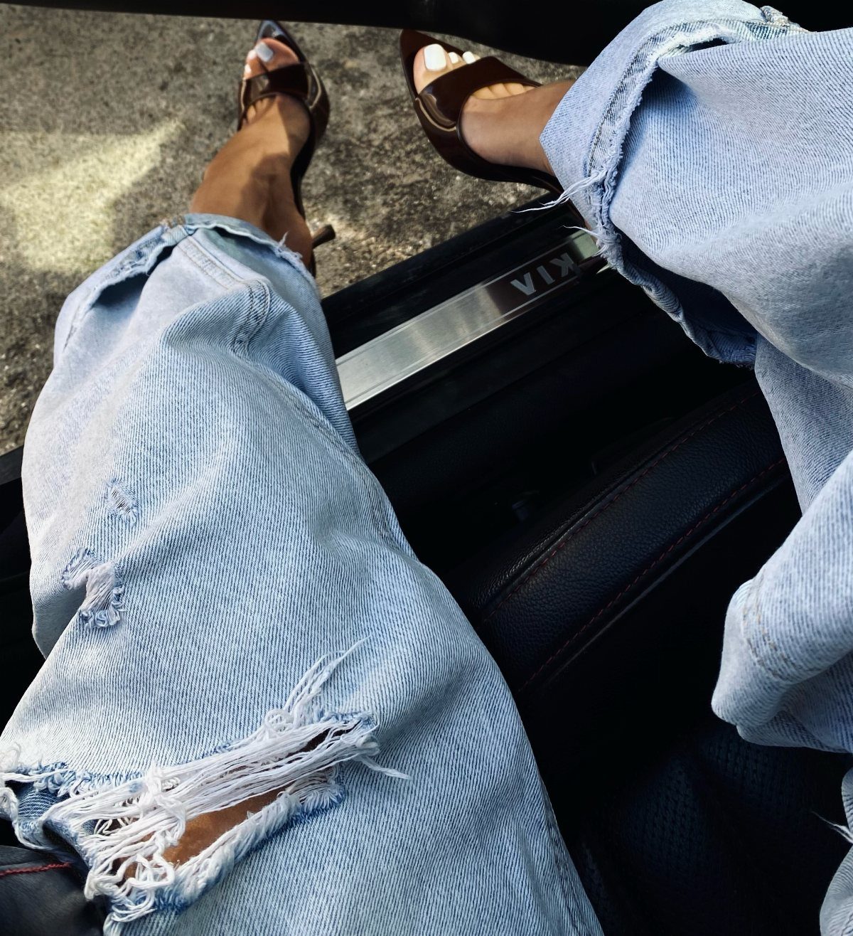 Person wearing light blue distressed denim jeans and brown kitten heel sandals while sitting in a car.