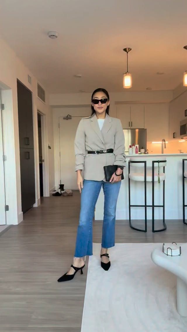 A woman posing in a beige blazer, blue flared jeans, and black kitten heels.