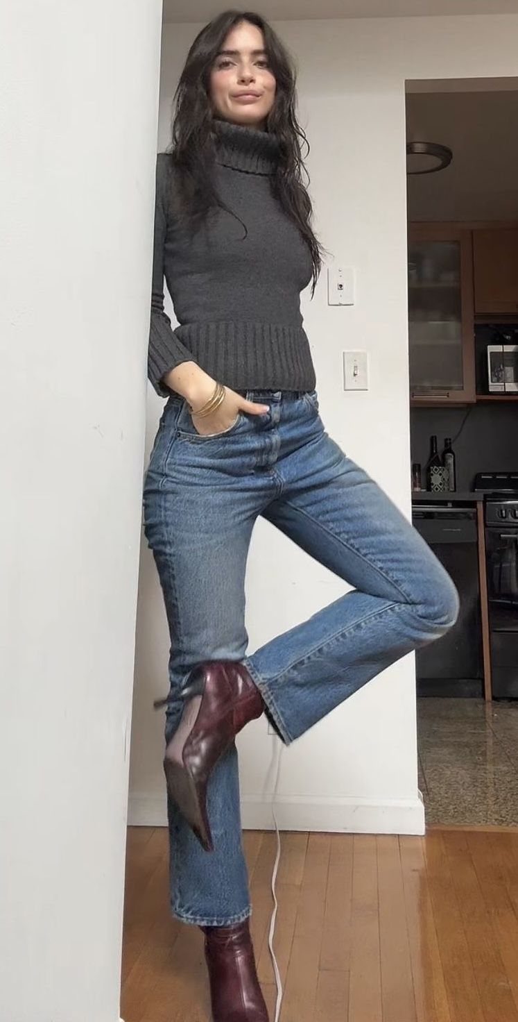 A woman posing in blue jeans, a grey turtleneck, and burgundy kitten heel boots.