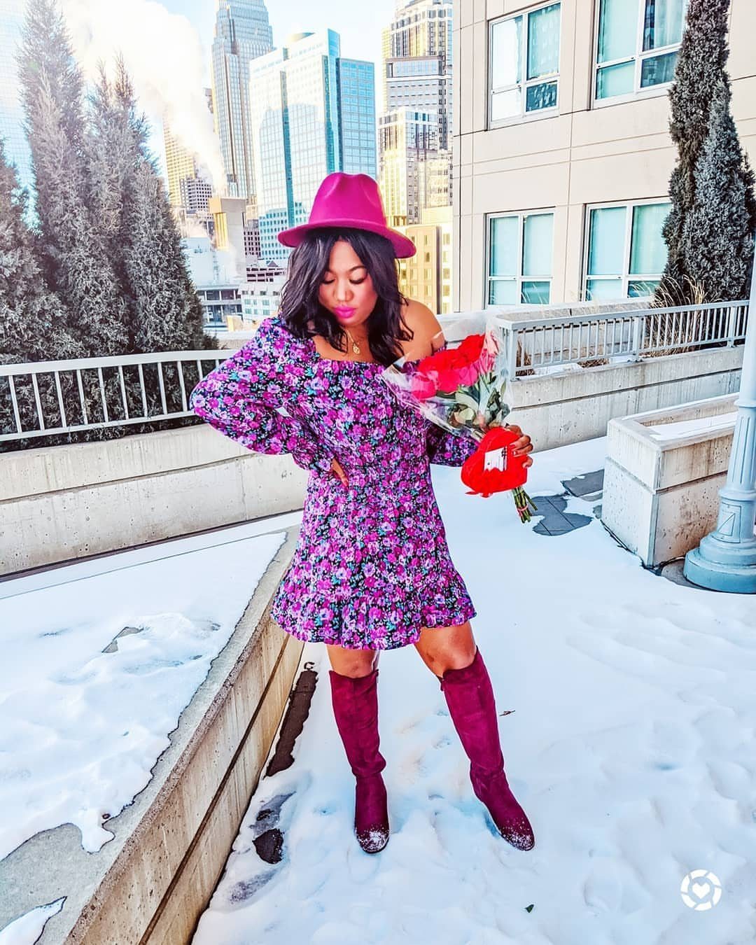 Woman in a purple floral mini dress and burgundy boots holding roses in the snow.