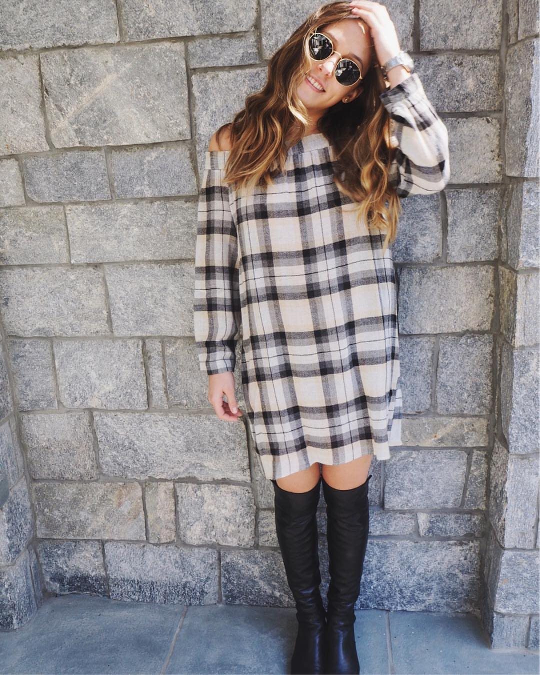 A woman wearing a plaid off-the-shoulder mini dress and black knee-high boots against a stone wall.