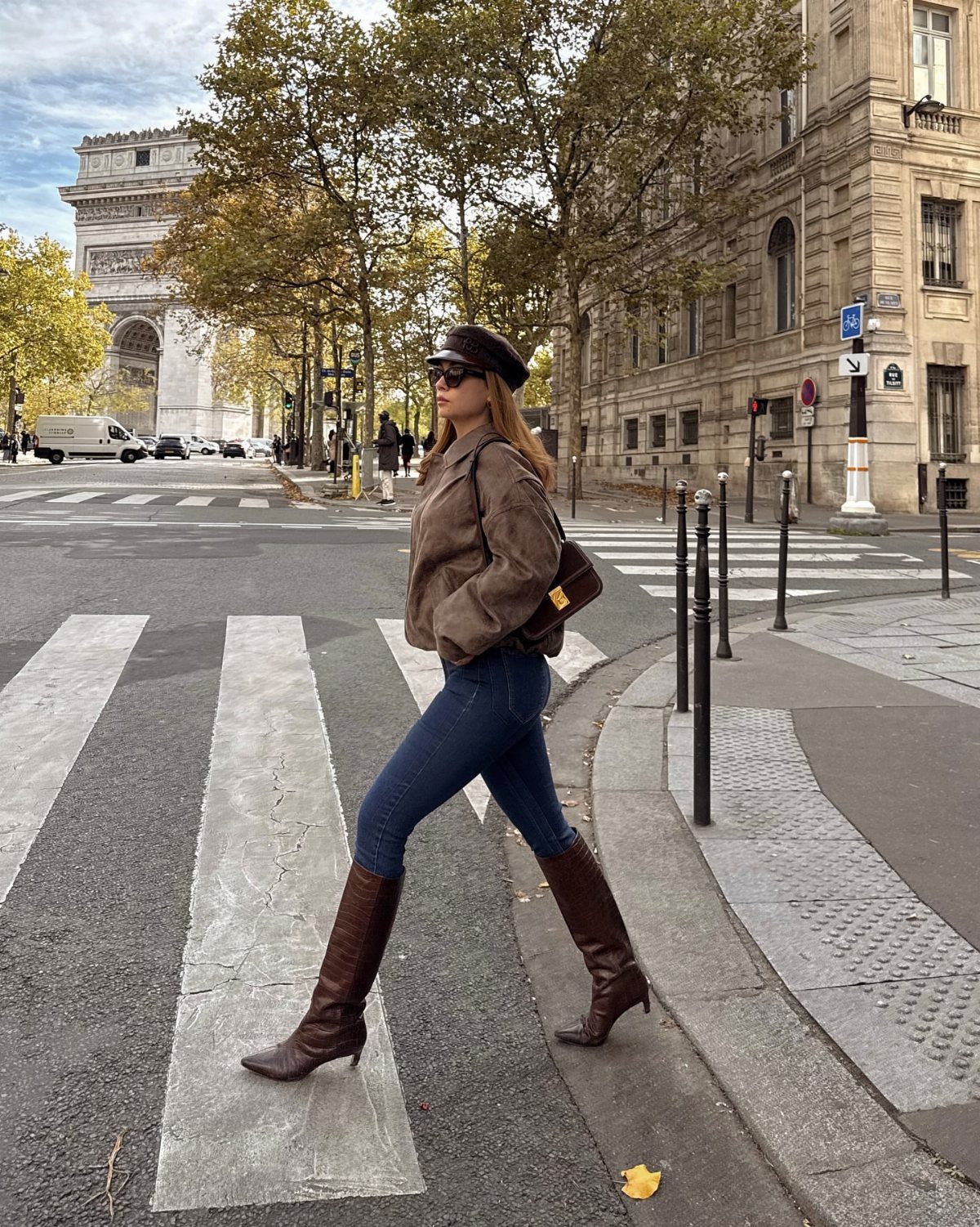 A woman in blue jeans and brown knee-high boots walking across a city street.