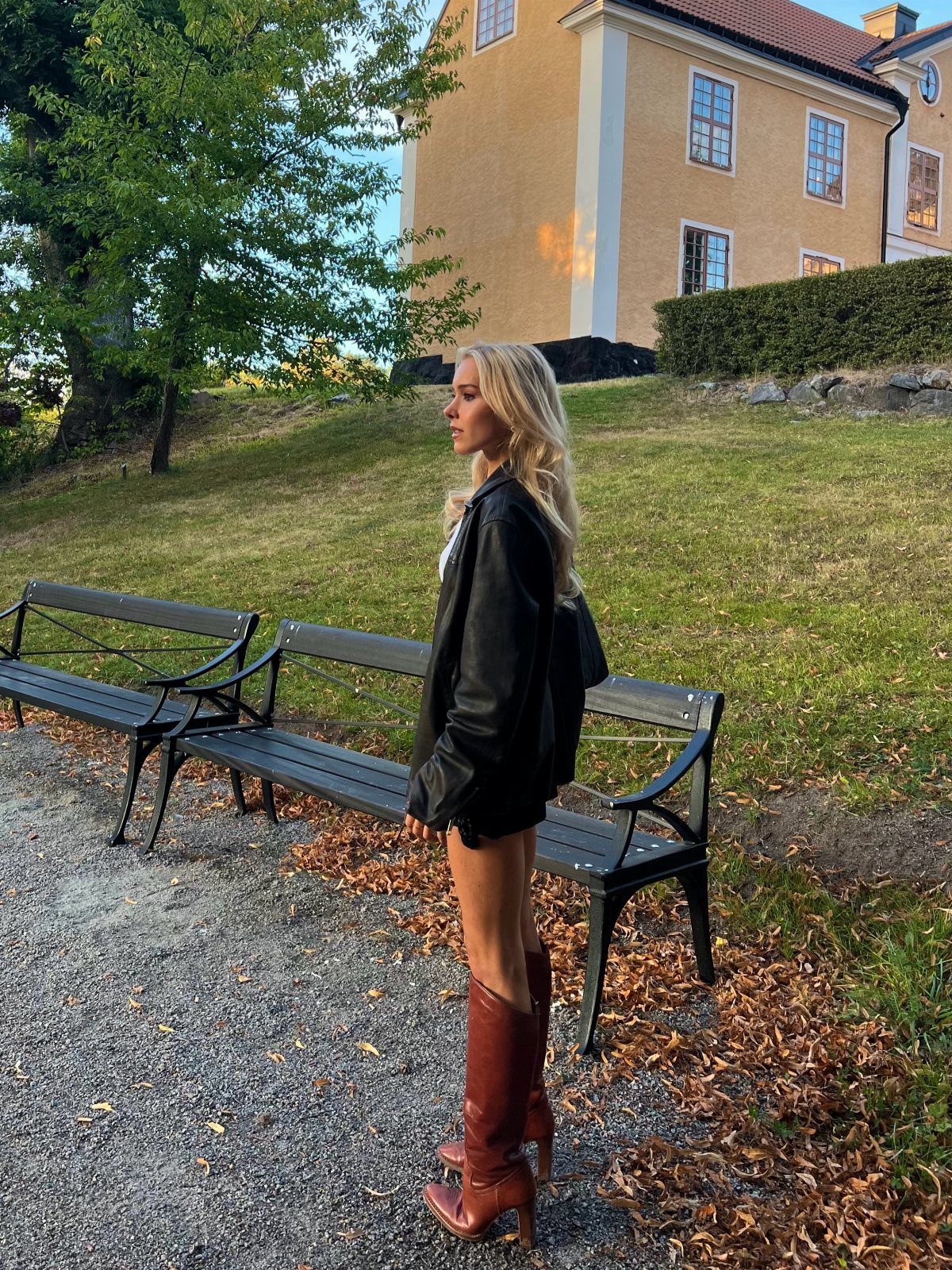 A blonde woman wearing a black leather jacket and brown knee high boots standing near a bench.