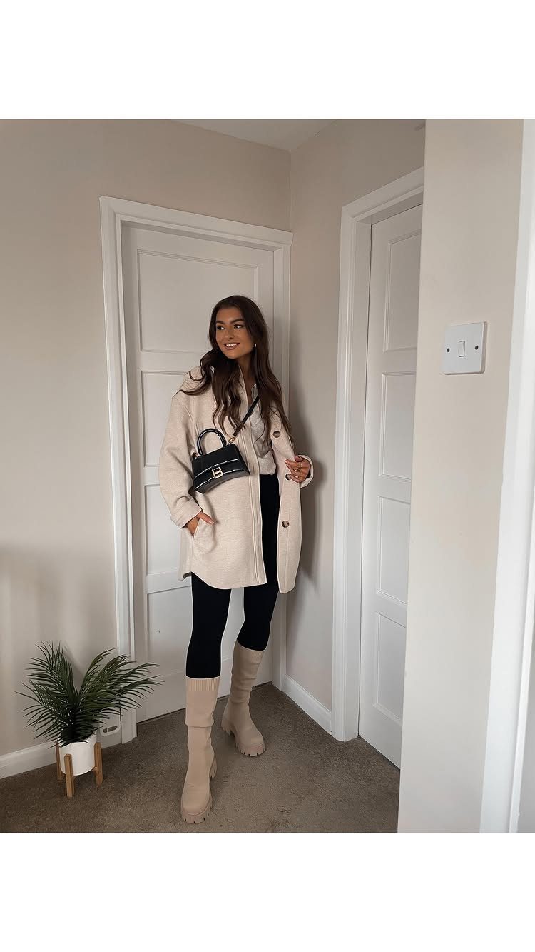 A woman wearing a cream coat, black leggings, and beige knee-high boots standing indoors.