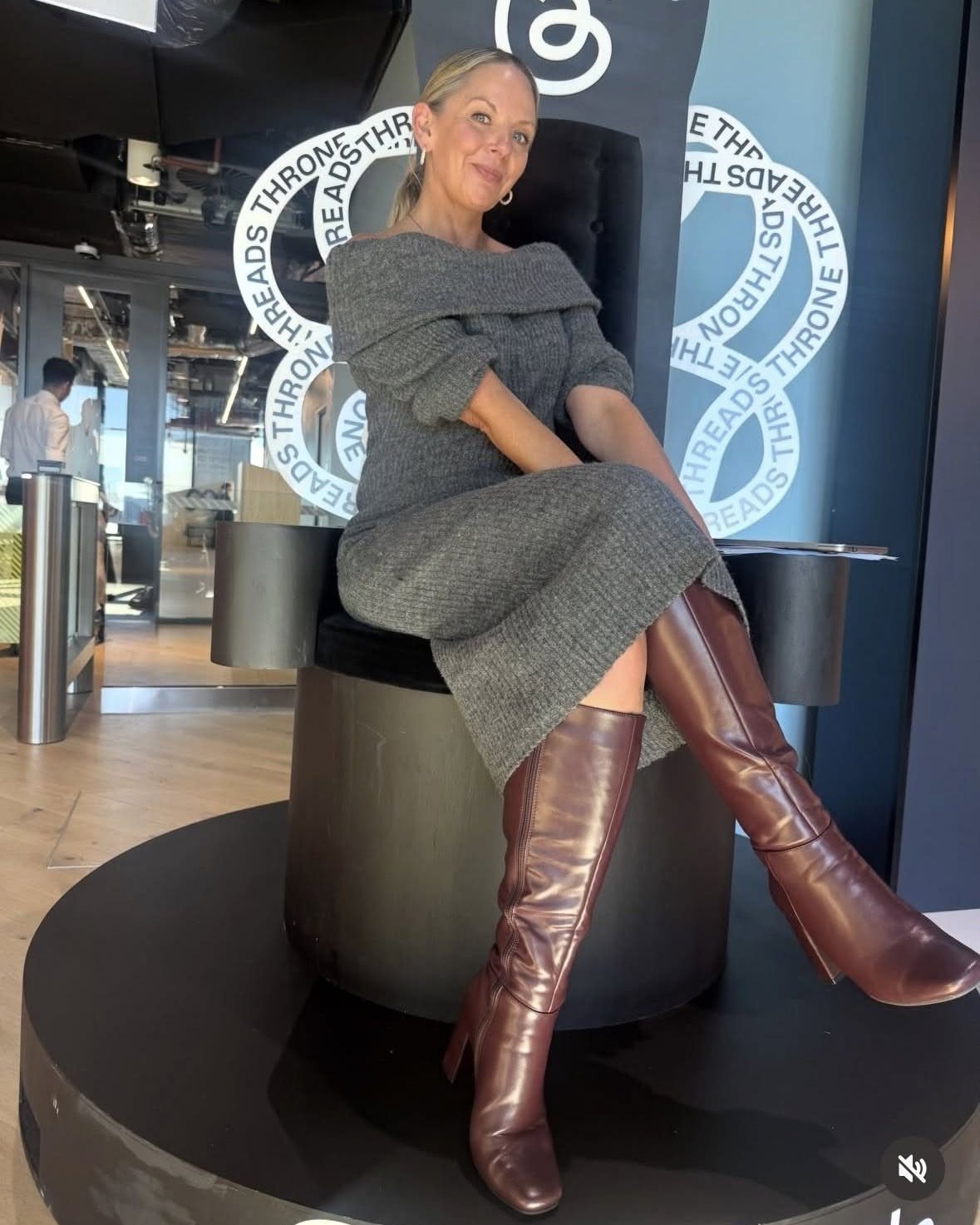 A woman wearing a grey sweater midi dress and reddish-brown knee-high leather boots sitting in a chair.