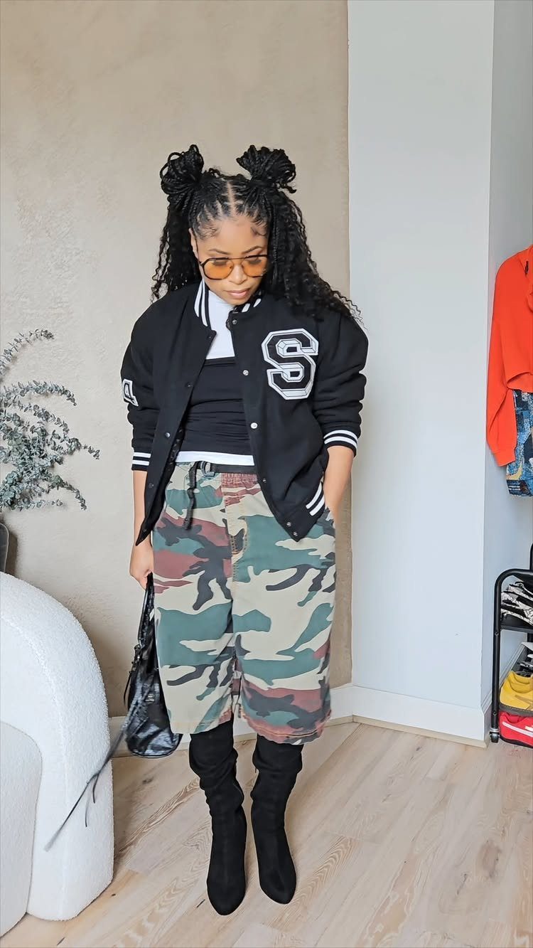 A woman posing in a black varsity jacket, camouflage shorts, and black knee-high boots.