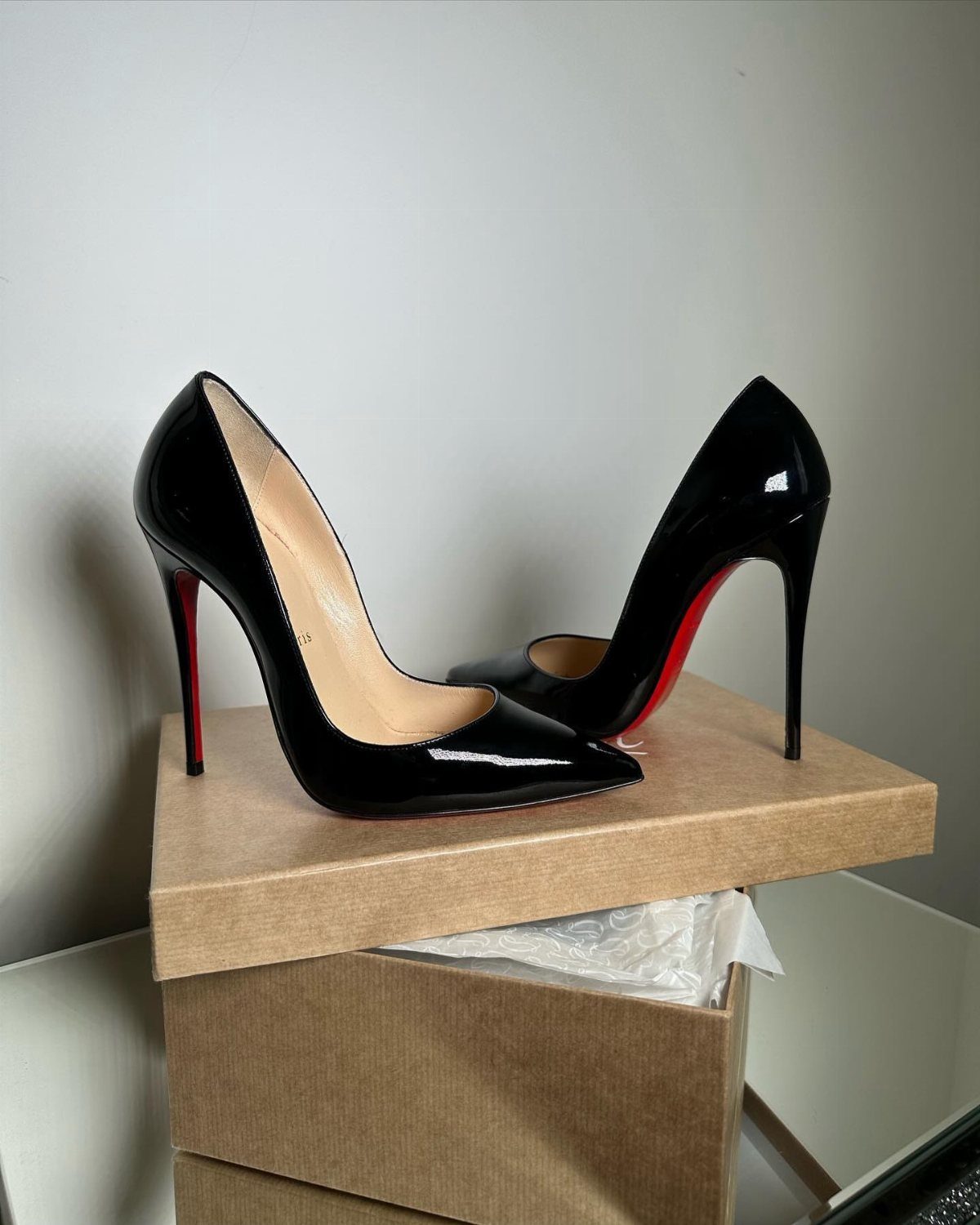 Black patent leather high-heeled pumps with distinctive red soles resting on a cardboard box.