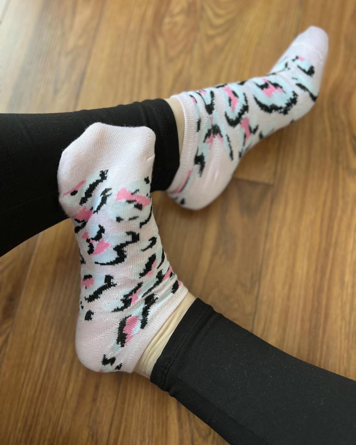 Person wearing patterned pink and black ankle socks with black leggings on a wooden floor.