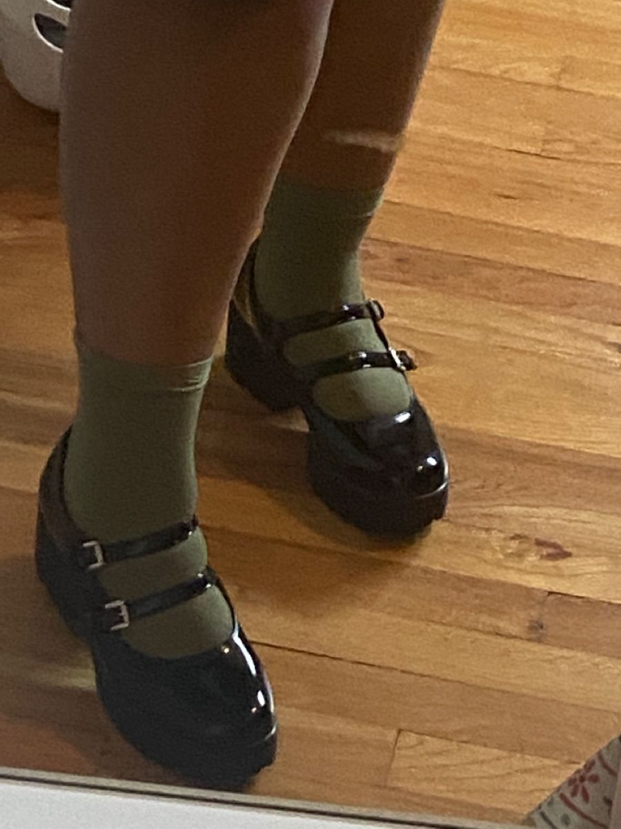 Person wearing olive green socks and black patent leather Mary Jane shoes on a wooden floor.