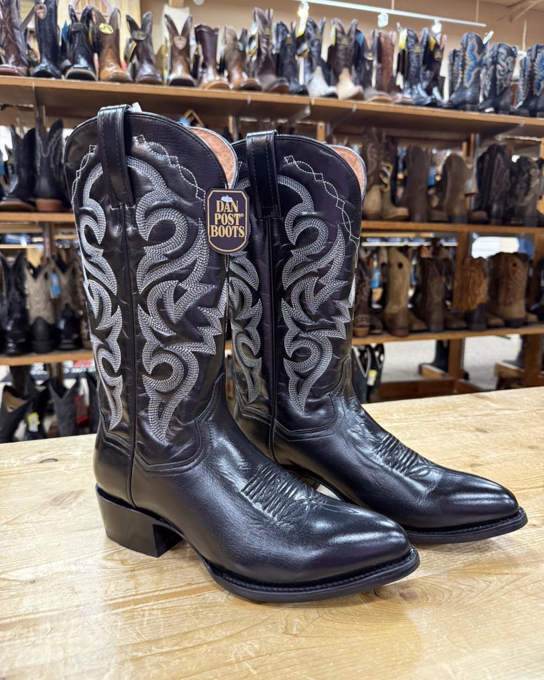 Black leather cowboy boots with silver stitching sitting on a wooden counter in a boot store.