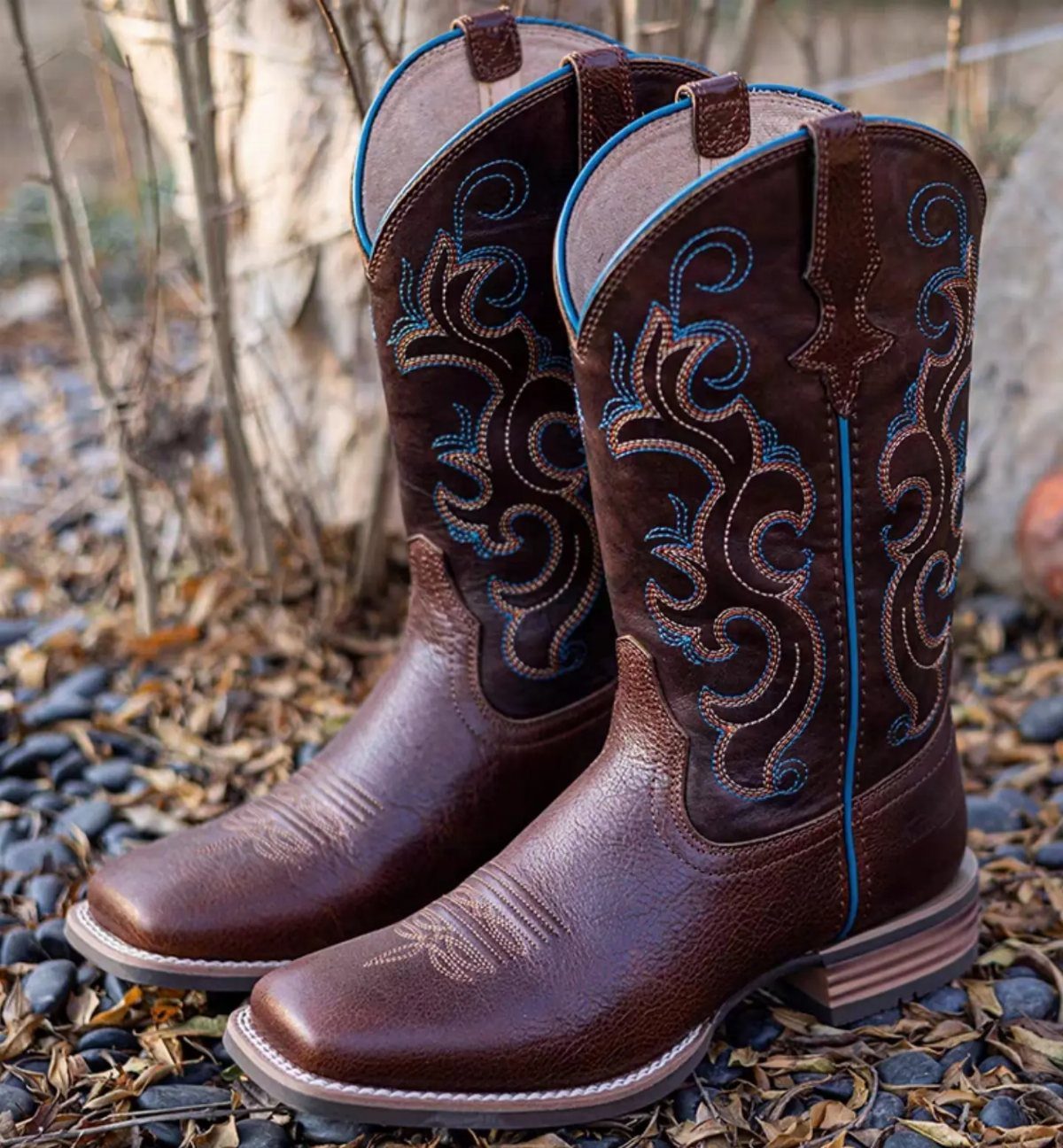 Brown leather western boots with blue stitching sitting on a bed of small grey pebbles.