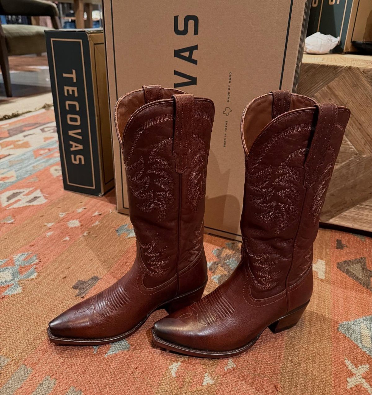 Brown leather cowboy boots standing on a patterned rug in front of a Tecovas box.