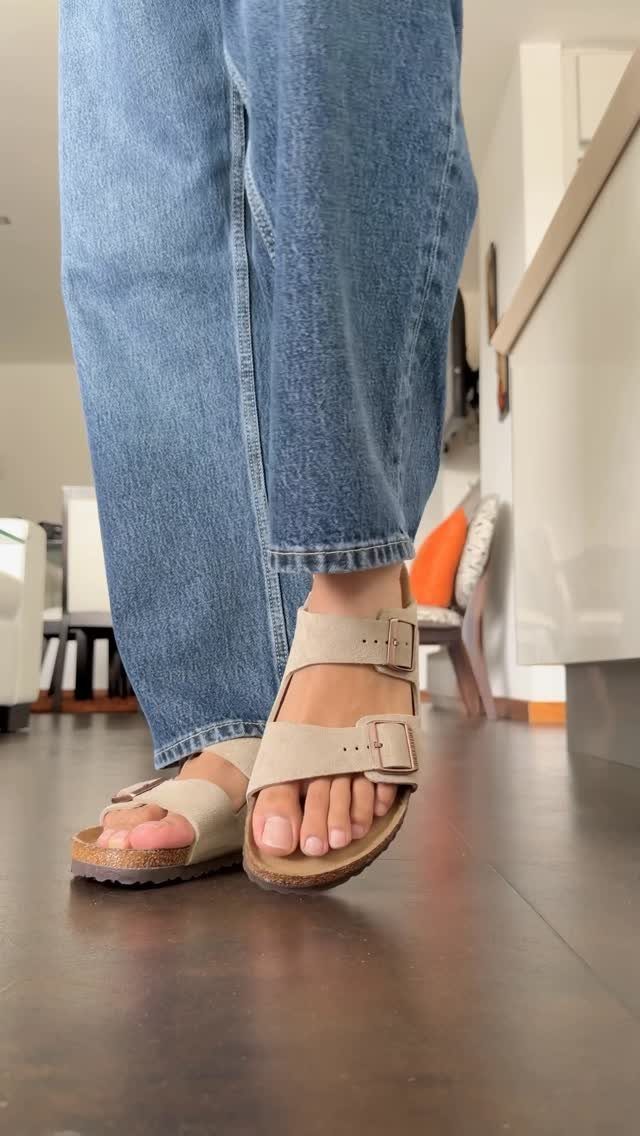 Person wearing blue jeans and beige two-strap Birkenstock sandals walking on a dark floor.