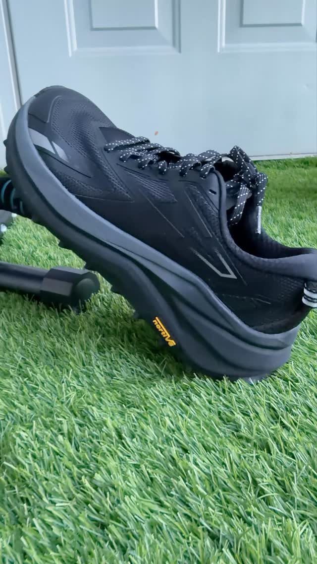 Black athletic walking shoe with speckled laces resting on green artificial turf.