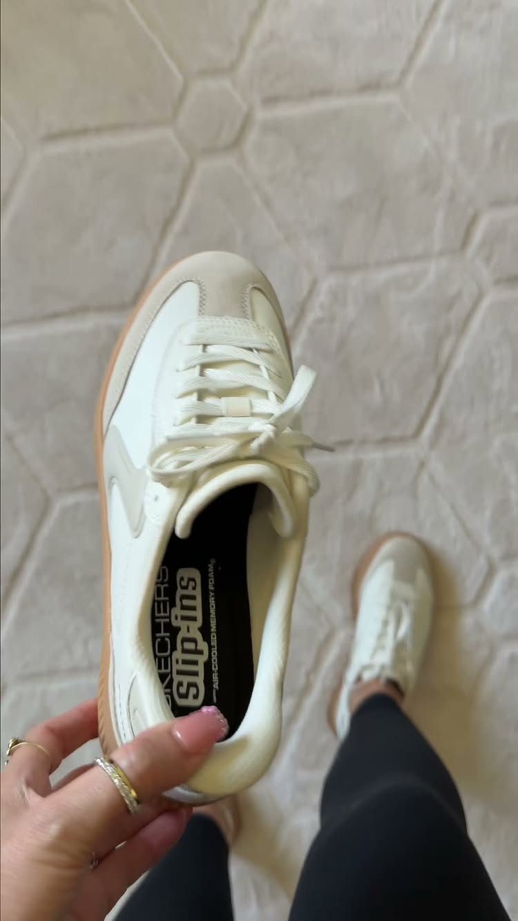 A person holding a white Skechers Slip-ins sneaker over their feet on a textured rug.