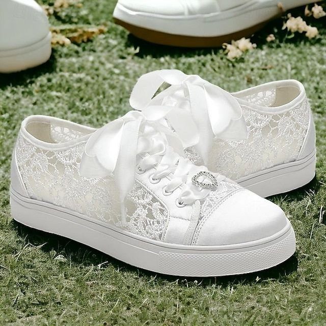 White lace wedding sneakers with large satin bows resting on green grass.