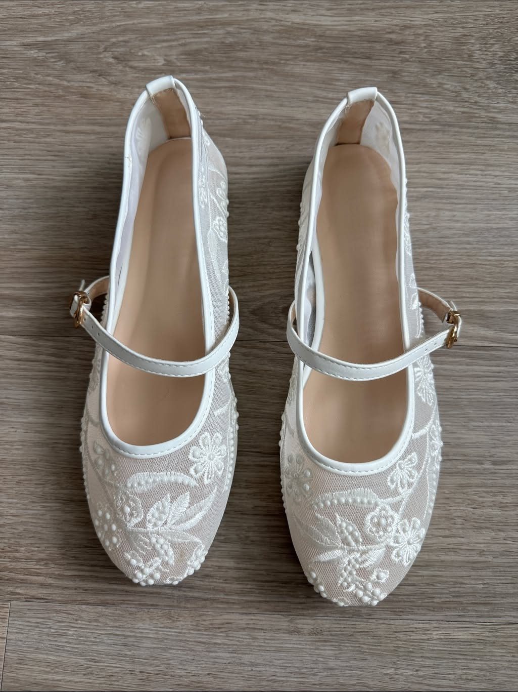 White embroidered wedding flats with pearl details and thin straps on a wooden floor.