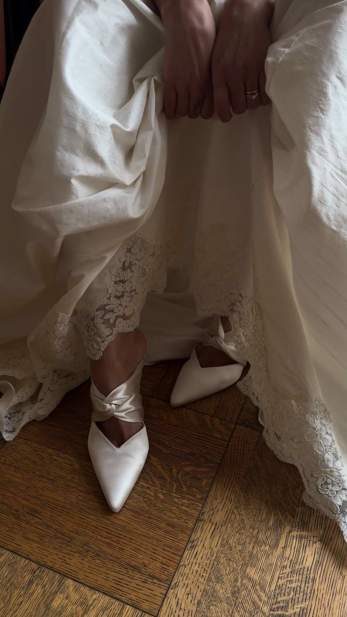 White satin pointed-toe wedding shoes with knotted straps worn under a lace wedding dress.