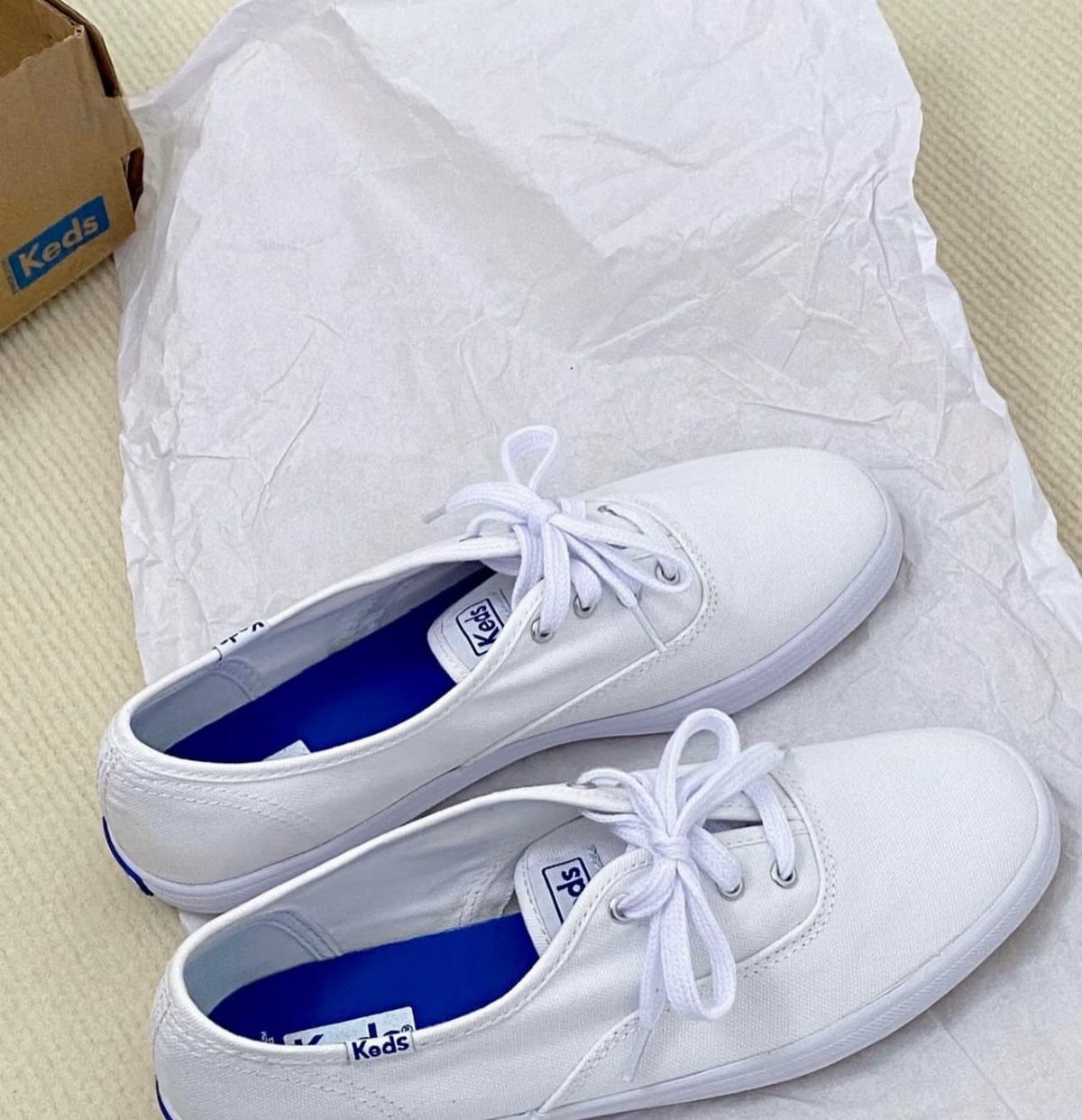 White Keds canvas sneakers with blue insoles resting on white crumpled tissue paper.