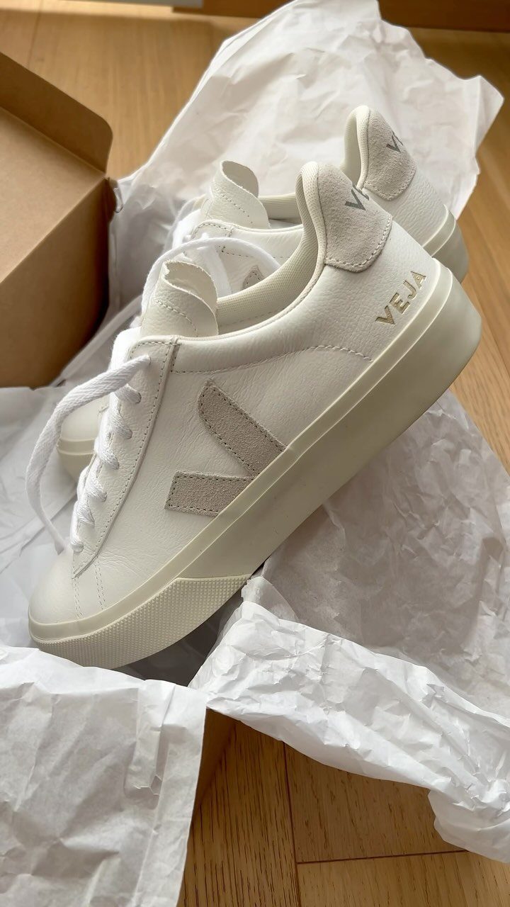 White Veja sneakers resting inside a cardboard box with white tissue paper on a wooden floor.