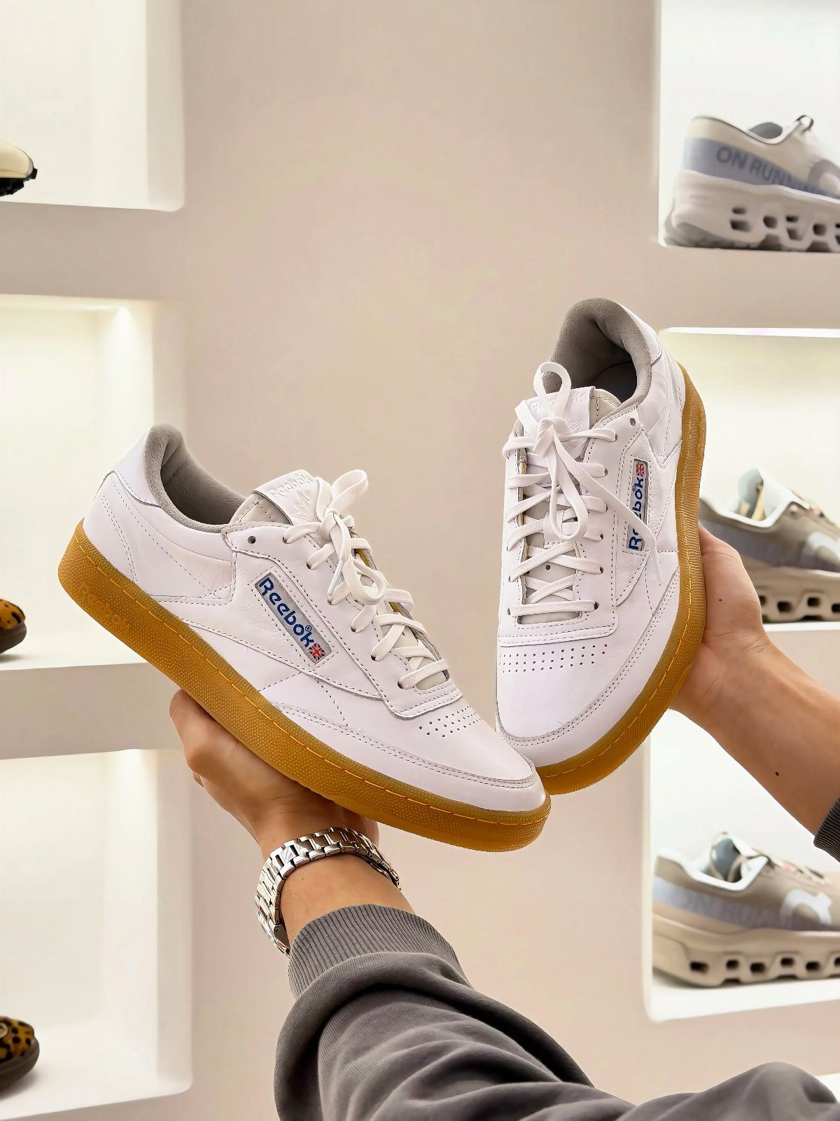 Hands holding up a pair of white Reebok sneakers with tan gum soles in a store.