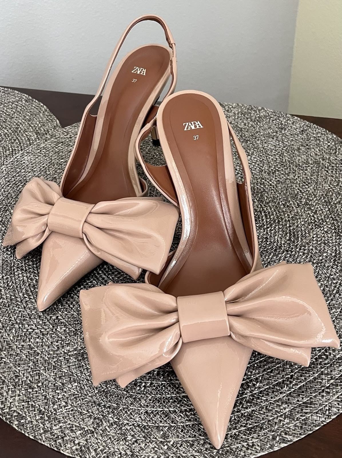 Nude pointed-toe high heel sandals with large decorative bows on a textured grey surface.