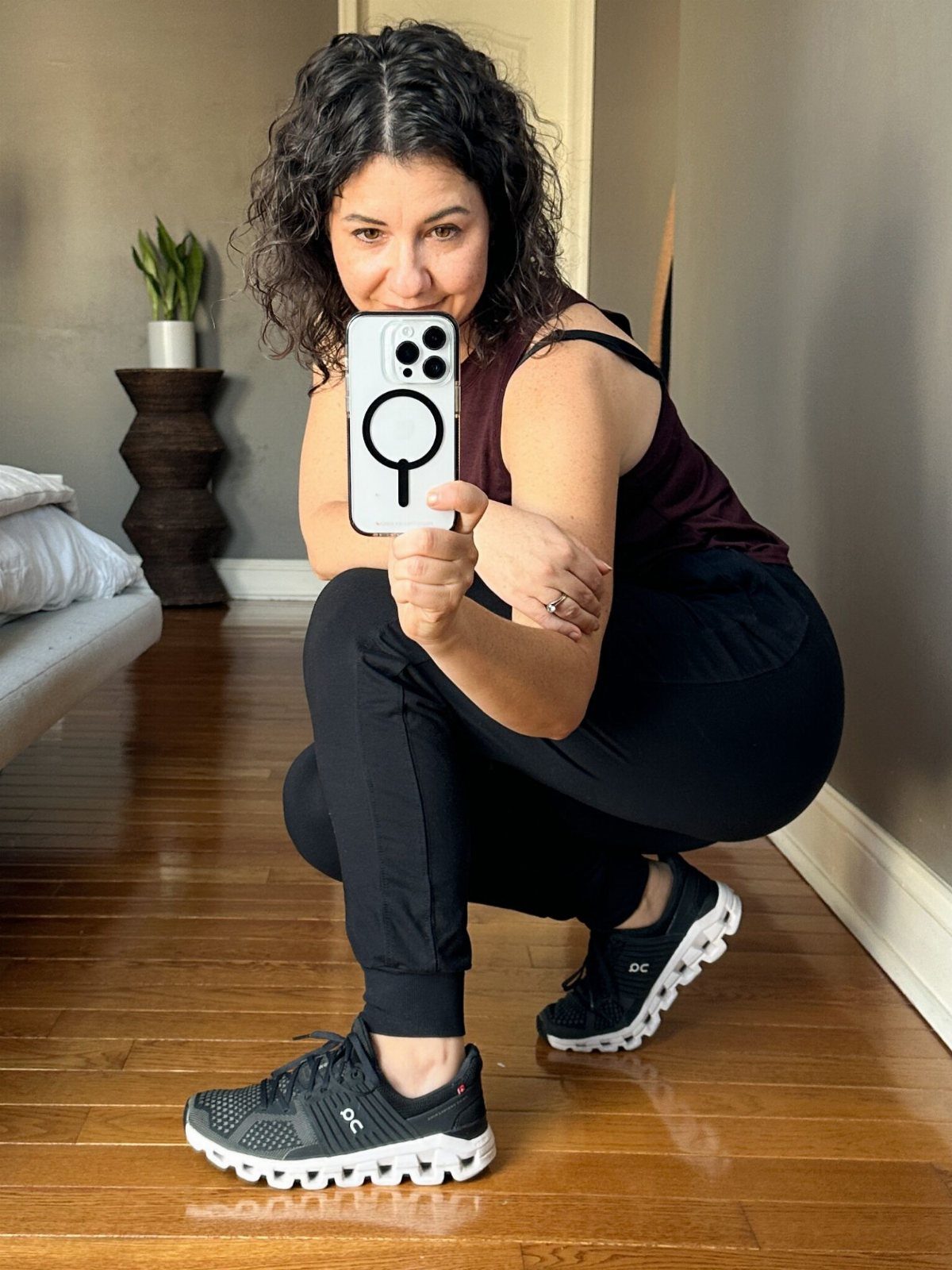 A woman in black athletic wear squats on a wooden floor wearing black On Cloud shoes.