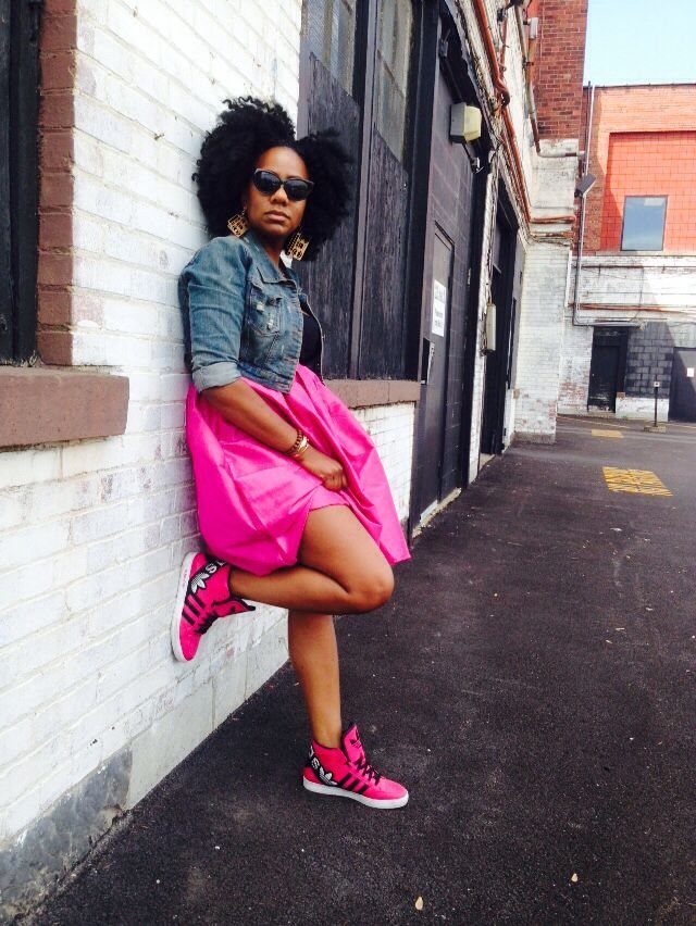 A woman in a denim jacket and pink skirt posing in an alley wearing pink sneakers.