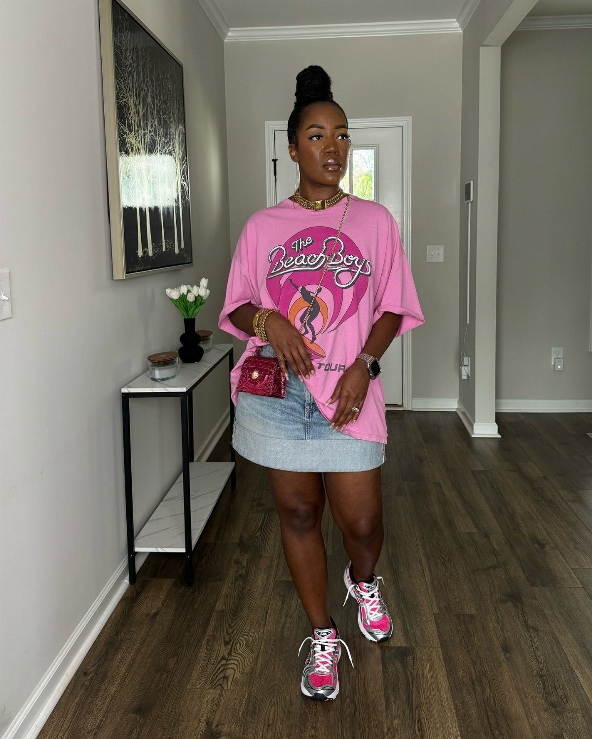 Woman wearing a pink Beach Boys graphic t-shirt, denim skirt, and pink sneakers in a hallway.