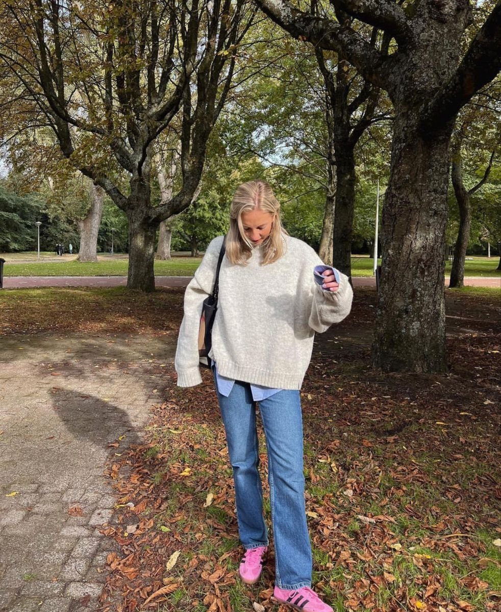 A woman wearing a cream oversized sweater, blue jeans, and pink sneakers standing in a park.