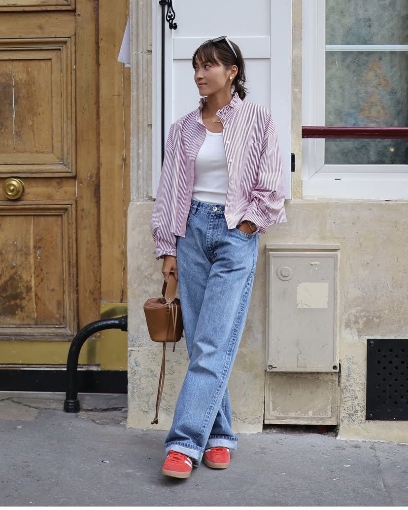 Woman wearing a pink striped button-down shirt, blue jeans, and pink sneakers standing by a door.