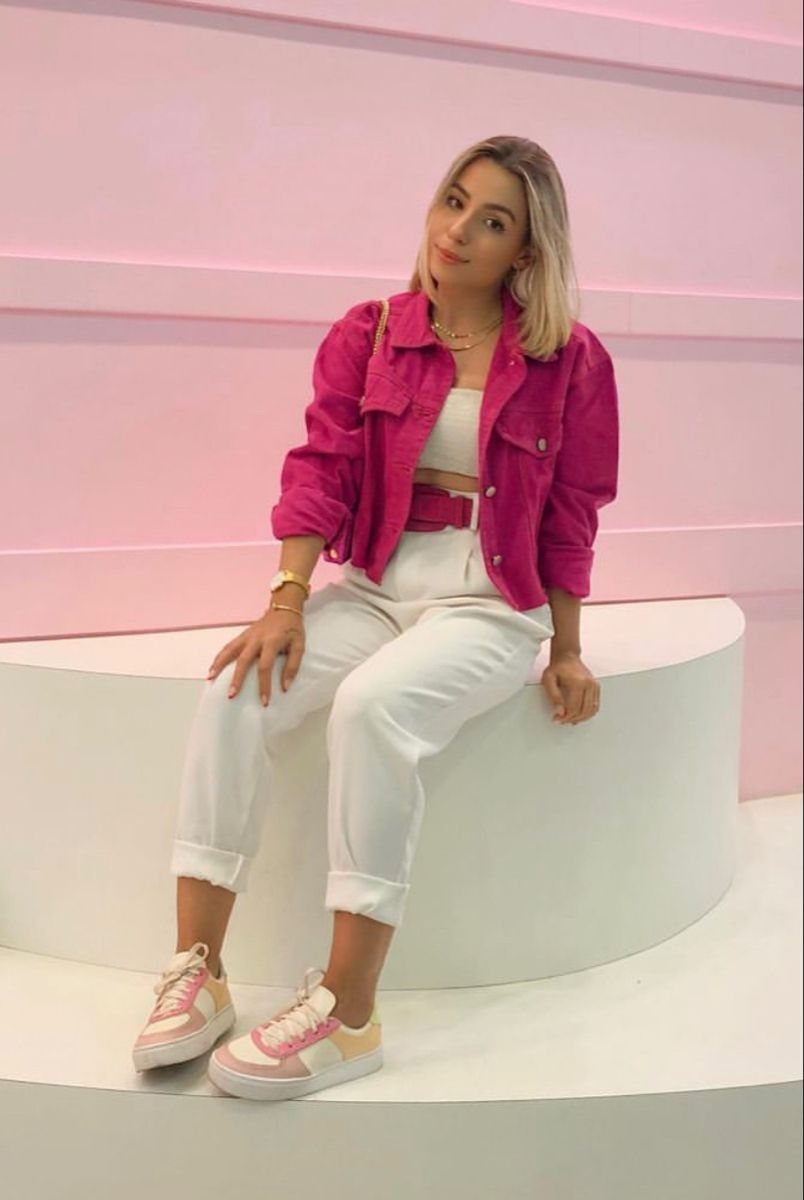 A woman wearing a pink cropped jacket, white top, white pants, and pink sneakers sitting on a bench.