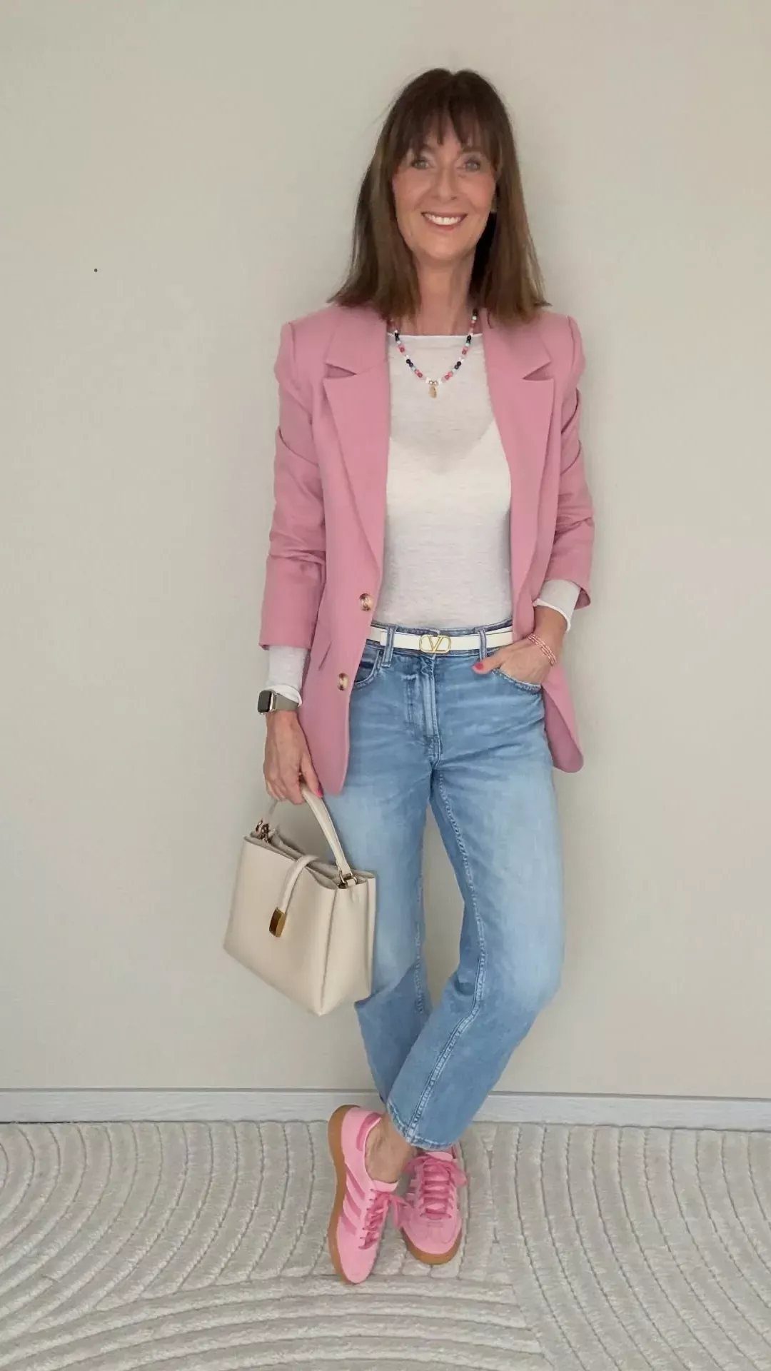 A woman wearing a pink blazer, white top, blue jeans, and pink sneakers standing against a wall.