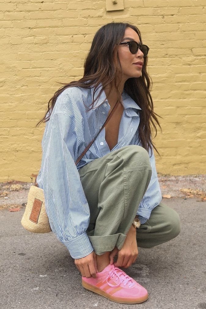 Woman crouching in olive trousers and a blue striped shirt wearing pink sneakers.