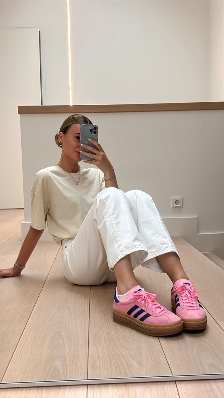 Woman in white pants and cream t-shirt wearing bright pink sneakers sitting on floor.