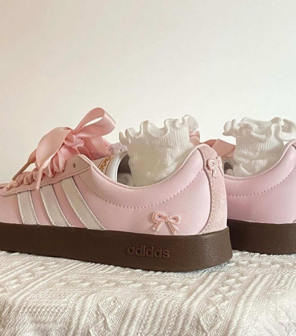 Light pink Adidas sneakers with white stripes and decorative bows paired with white ruffled socks.