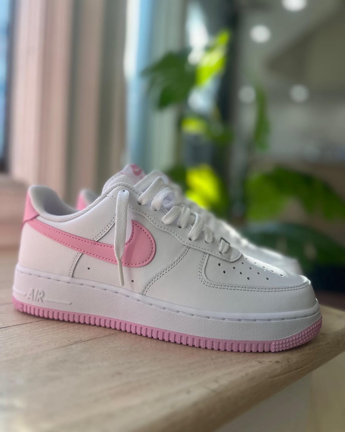 White Nike Air Force 1 sneakers with pink swooshes and pink soles on a wooden table.