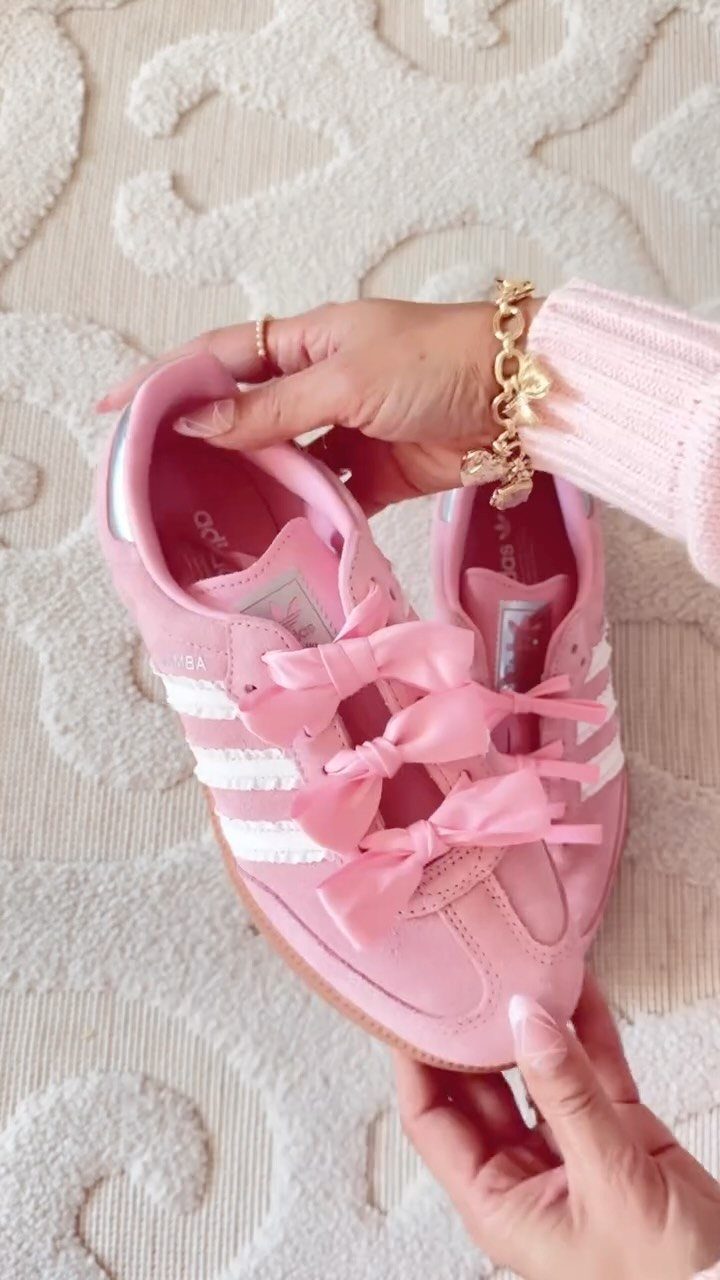 Hands holding a pair of pink Adidas sneakers decorated with large pink satin bows.