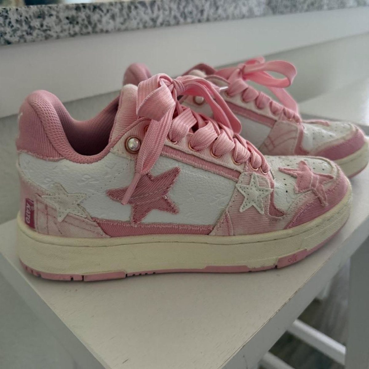 Pink and white casual sneakers featuring star appliqués and pink laces sitting on a white shelf.