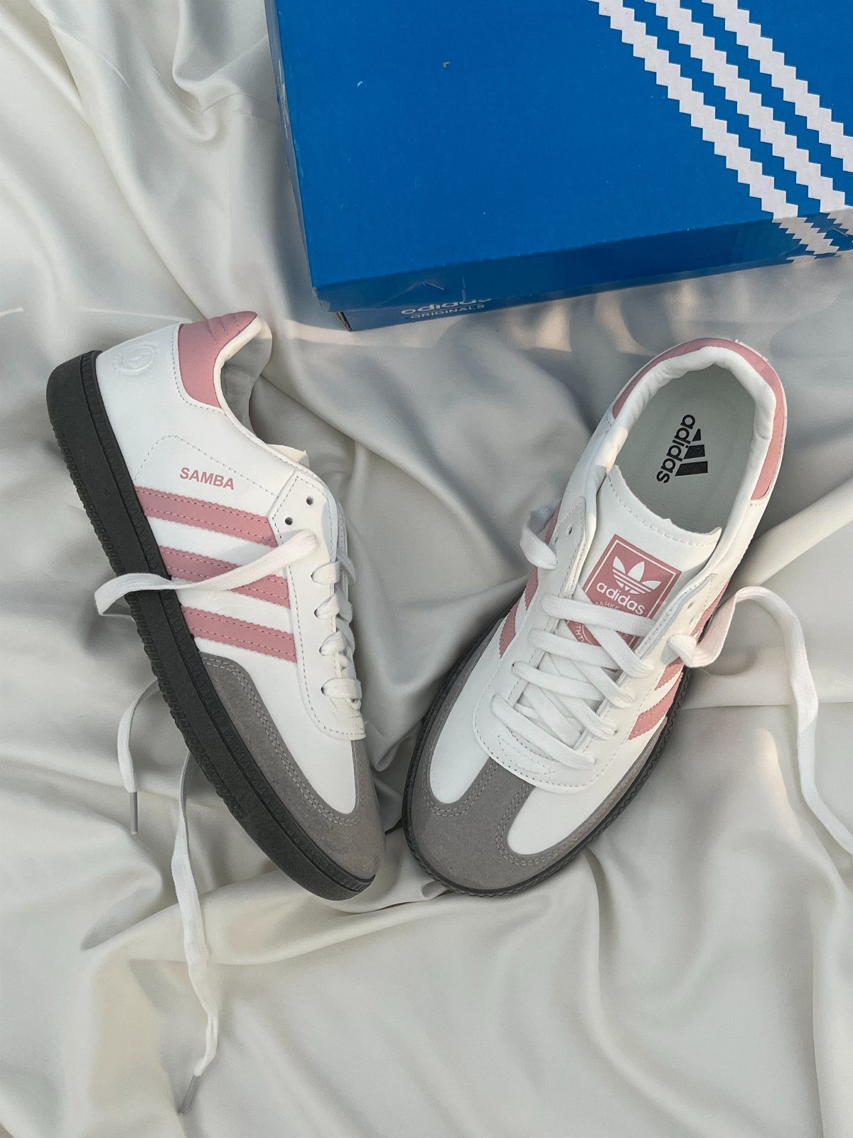 White and pink Adidas Samba sneakers resting on white fabric next to a blue shoebox.